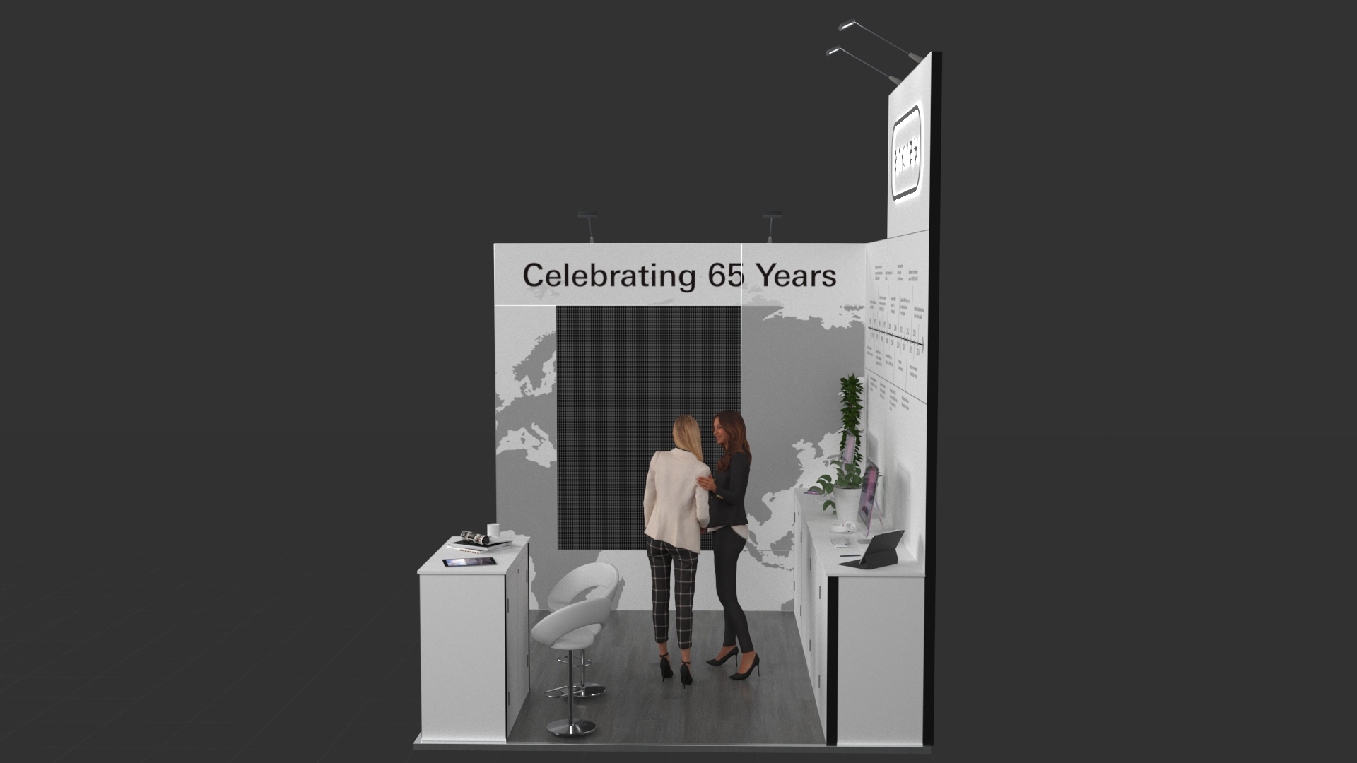 L-shaped exhibition stand featuring a reception counter and two workstations with computer monitors and under-counter storage. The stand has an elevated central back panel and large wall graphics.