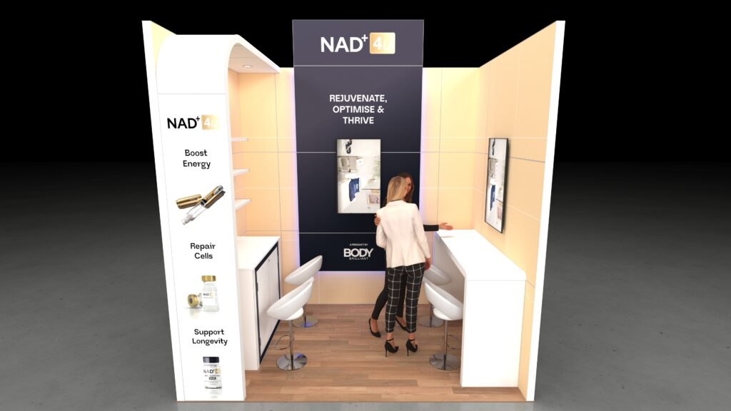 U-shaped exhibition stand featuring a central back wall with an elevated header panel. The left side has a vertical display column with product shelving, and the right side is an extended high-top counter/bar with two stools. Two people are interacting at the stand.