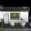 3D rendering of an exhibition stand design featuring a front reception counter, a central meeting area with four round lounge chairs, built-in wall display shelves, a small refreshment station, and a closed door leading to a private storage room.